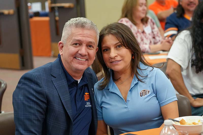 Individual wearing a UTEP Engineering polo shirt speaks from a podium during a homecoming recognition ceremony, with a large presentation slide displaying an alumni portrait visible behind the speaker and College of Engineering balloon decorations positioned beside the stage.